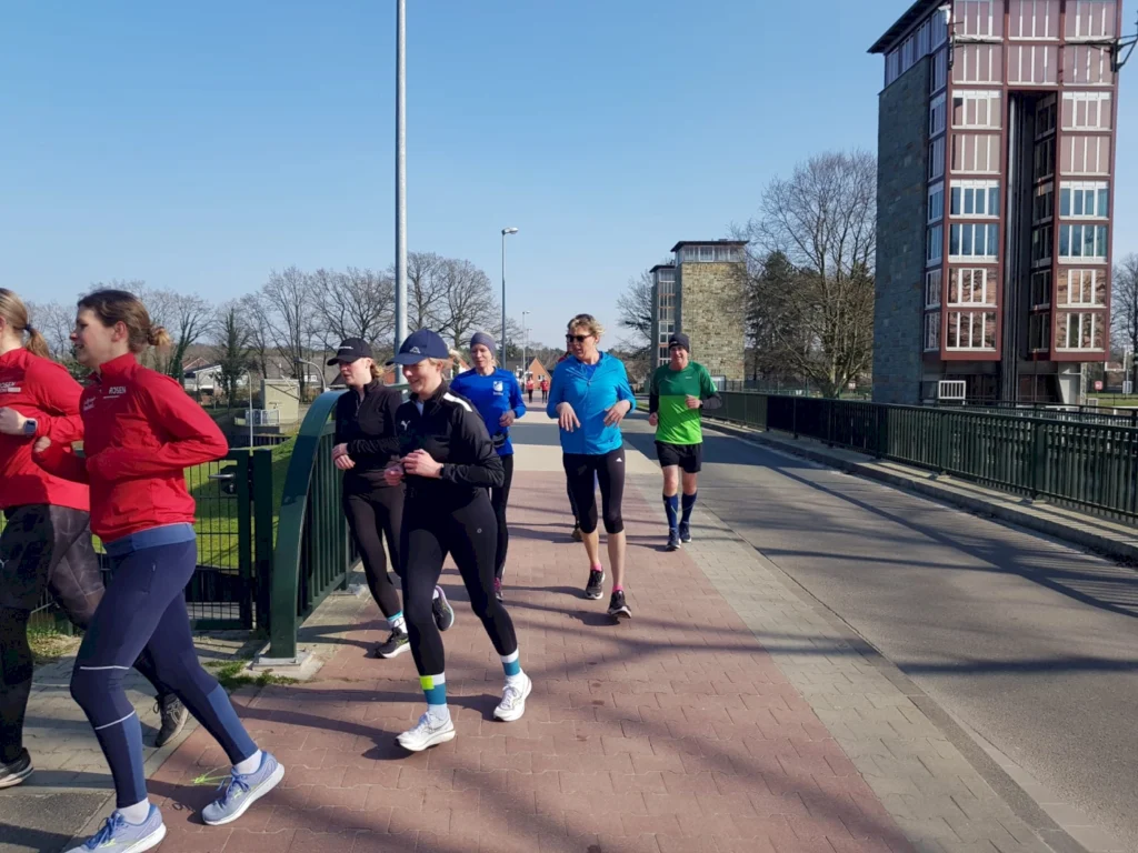 Gruppe von Läufern in Sportkleidung joggt auf einer Brücke an einem sonnigen Tag, umgeben von Bäumen und modernen Gebäuden.
