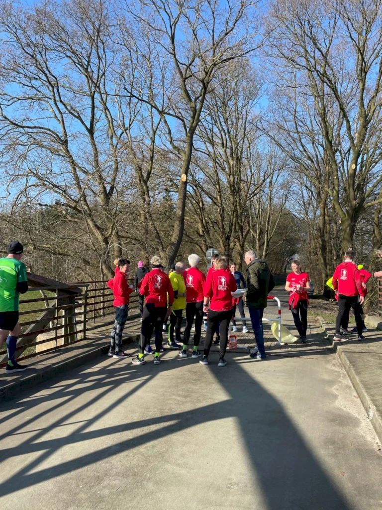 Gruppe von Läufern in roten T-Shirts versammelt sich auf einer Brücke in einem Park bei sonnigem Wetter. Bäume ohne Blätter im Hintergrund, symbolisiert die aktive Freizeitgestaltung und den Gemeinschaftssport.