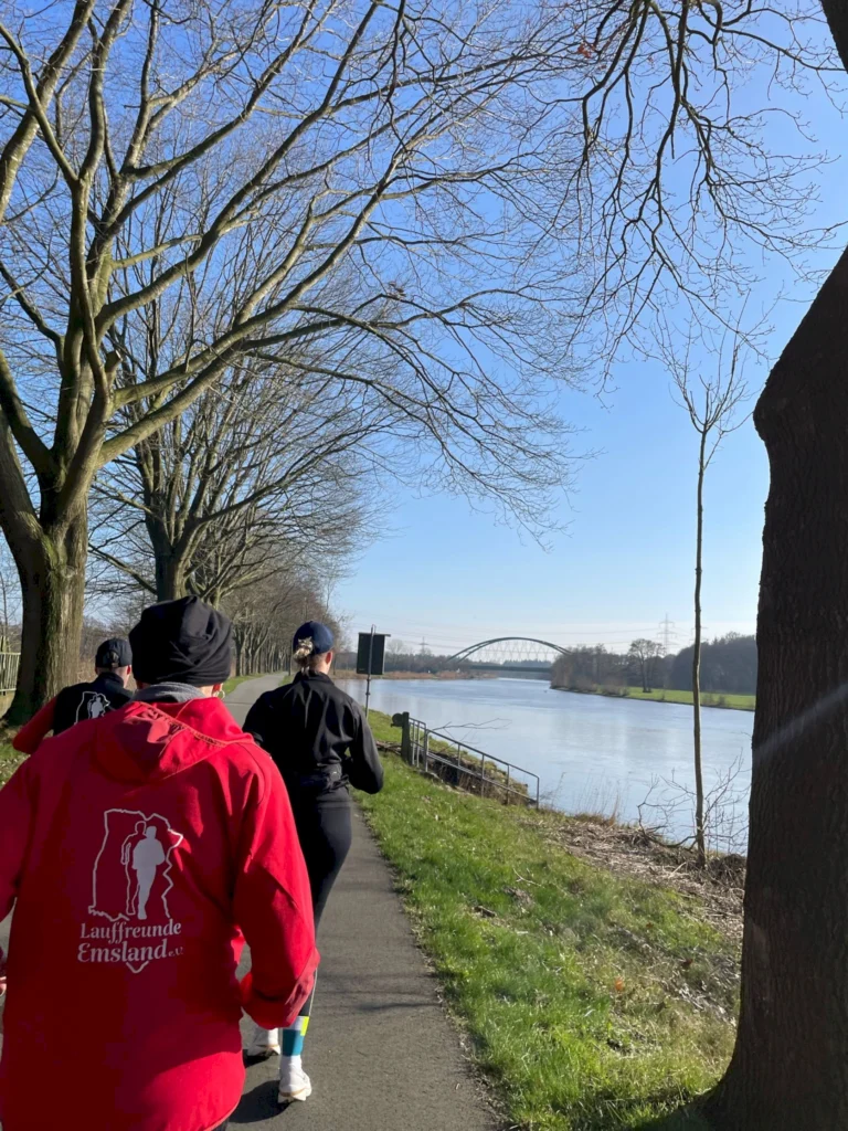 Läufergruppe am Ufer eines Flusses in Emsland, umgeben von Bäumen und klarem Himmel, ideal für Outdoor-Sportaktivitäten.