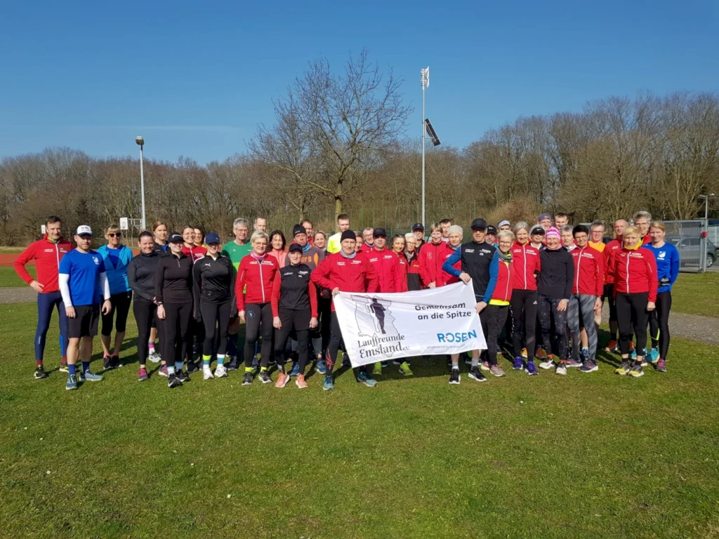 Gruppenfoto von Läufern und Sportlern im Freien, die in Sportkleidung gekleidet sind und eine Banner mit der Aufschrift „Gemeinsam an die Spitze“ halten. Die Szene zeigt einen sonnigen Tag auf einem Sportplatz mit Bäumen im Hintergrund.
