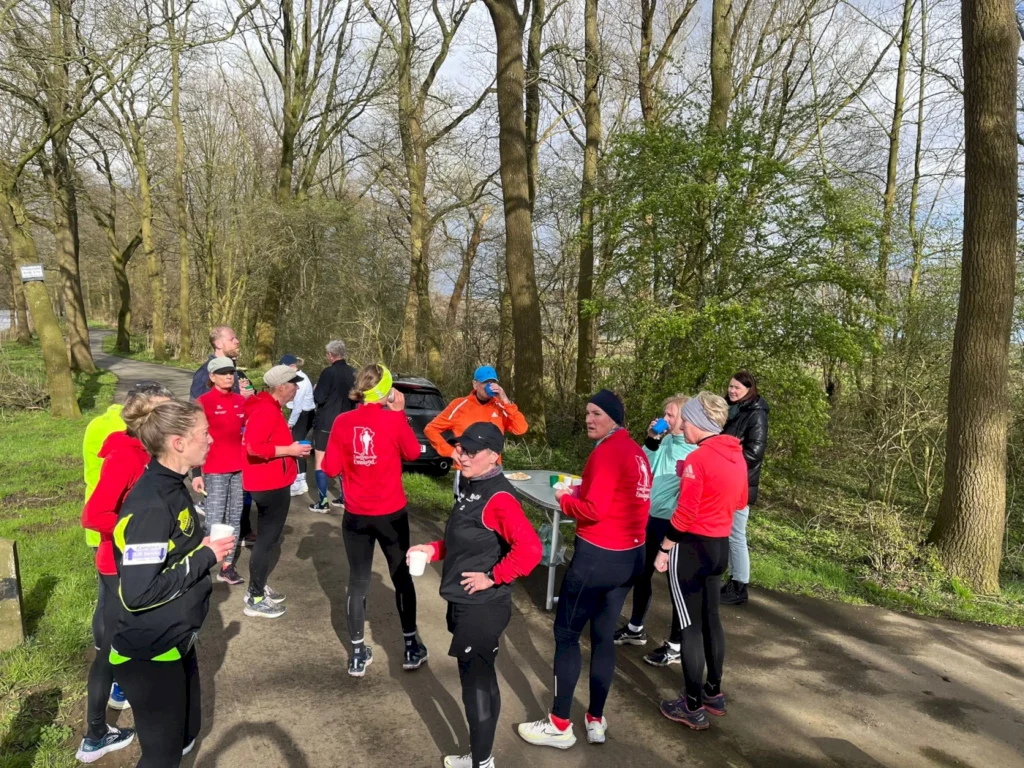Gruppe von Läufern in sportlicher Kleidung, die sich in einem Waldgebiet versammelt haben, während sie Getränke konsumieren und sich unterhalten. Im Hintergrund sind Bäume und ein Weg sichtbar, der in die Natur führt.