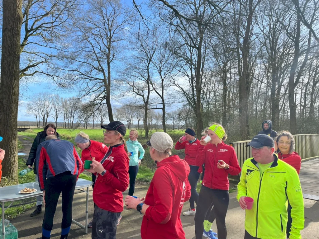 Gruppe von Läufern in roten Trainingsjacken bei einer Erfrischungsstation im Freien, umgeben von Bäumen und einer sonnigen Atmosphäre. Einige Teilnehmer genießen Snacks und Getränke, während andere sich unterhalten.