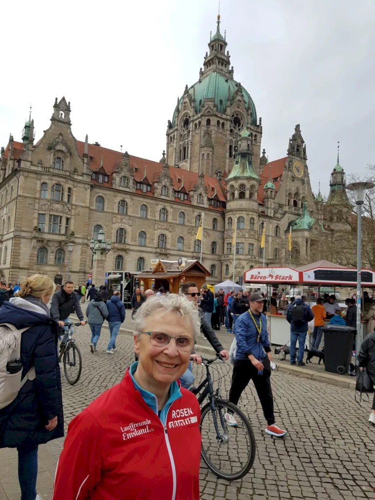 Frau mit grauen Haaren und Brille steht lächelnd vor dem historischen Rathaus von Hannover, umgeben von Menschen und Ständen auf einem belebten Platz.