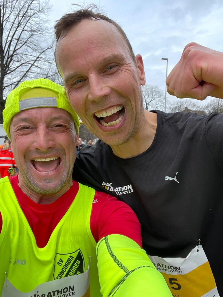 Lächelnde Läufer beim Marathon in Hannover, tragen sportliche Kleidung und Medaillen, im Hintergrund sind Zuschauer und Bäume zu sehen.