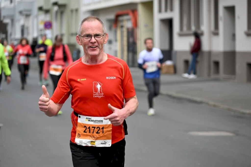 Läufer beim ADAC Marathon in Hannover, der in einem roten Shirt mit der Aufschrift "Rosen" fröhlich läuft und einen Daumen hoch zeigt. Im Hintergrund sind weitere Teilnehmer und die Straßenszene zu sehen.