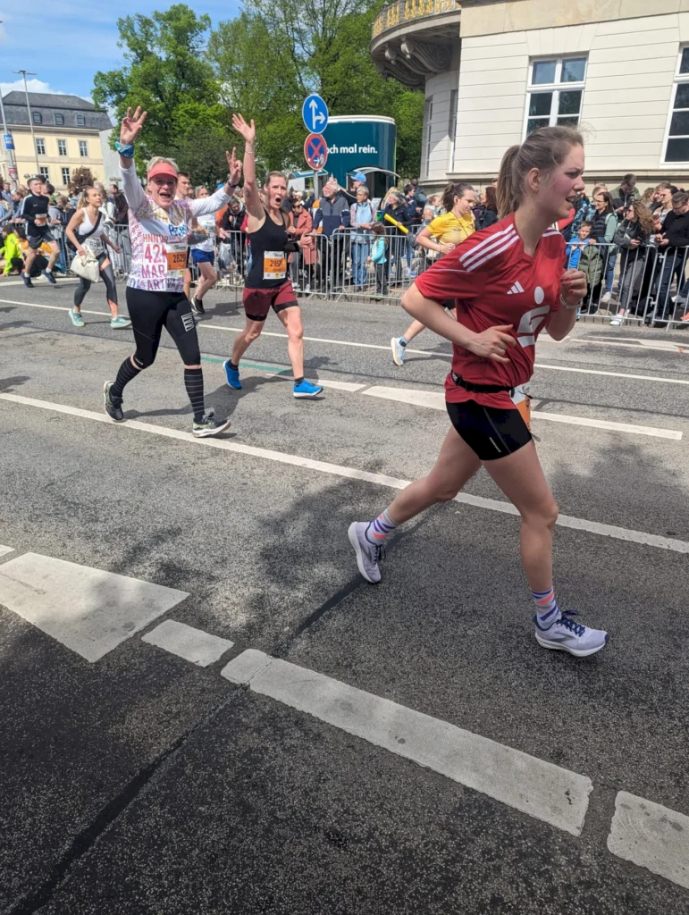 Teilnehmerinnen eines Straßenrennens laufen fröhlich und motiviert auf der Strecke, während Zuschauer sie anfeuern. Die Szene zeigt mehrere Läufer in sportlicher Kleidung und mit Startnummern, umgeben von Menschenmengen und einer festlichen Atmosphäre.