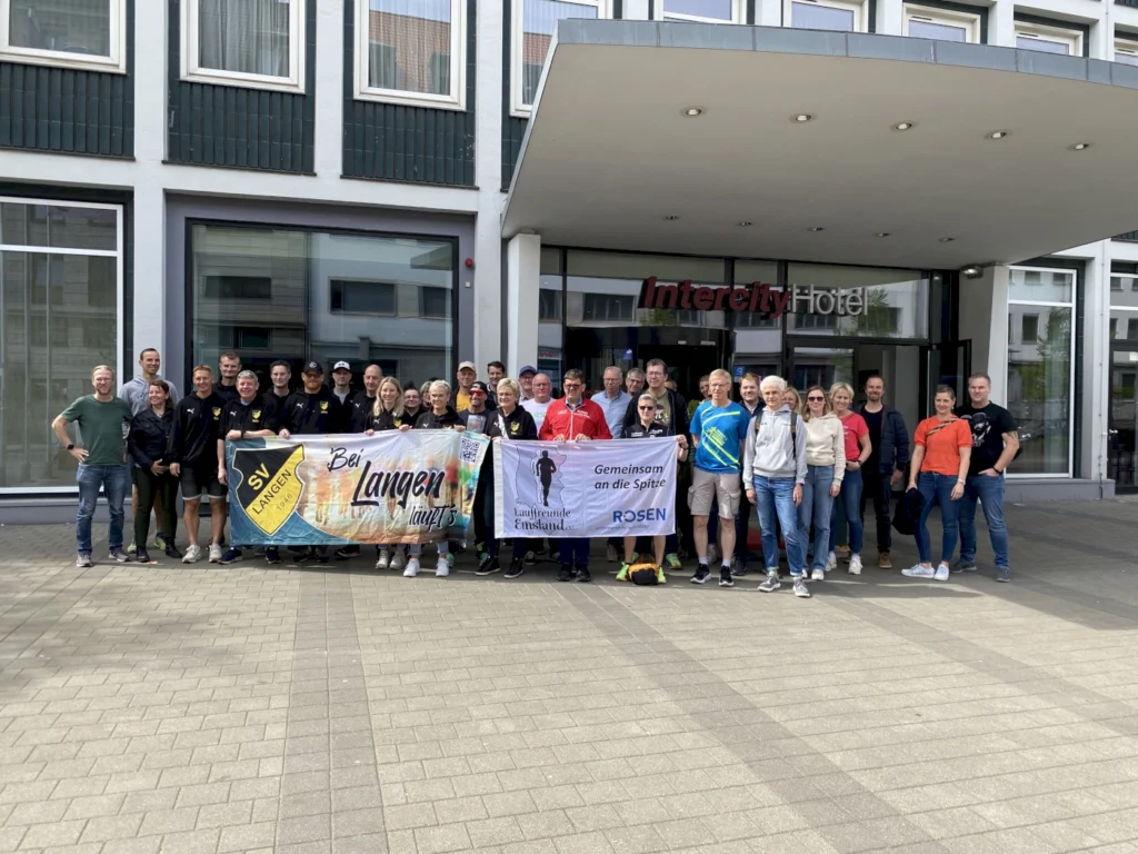 Gruppenfoto vor dem Intercity Hotel mit Teilnehmern des SV Langen und Laufgemeinschaft Emsland, die für ein gemeinsames Lauf-Event posieren. Banner mit den Aufschriften „Bei Langen läuft’s“ und „Gemeinsam an die Spitze“.