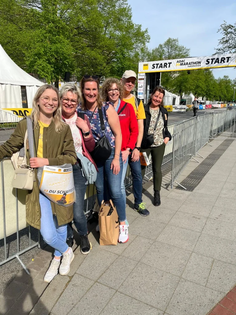 Sechs Personen stehen vor dem Startbereich des ADAC Marathons in Hannover, umgeben von grünen Bäumen und festlichen Zeltinstallationen. Sie lächeln und scheinen aufgeregt über das bevorstehende Event zu sein.
