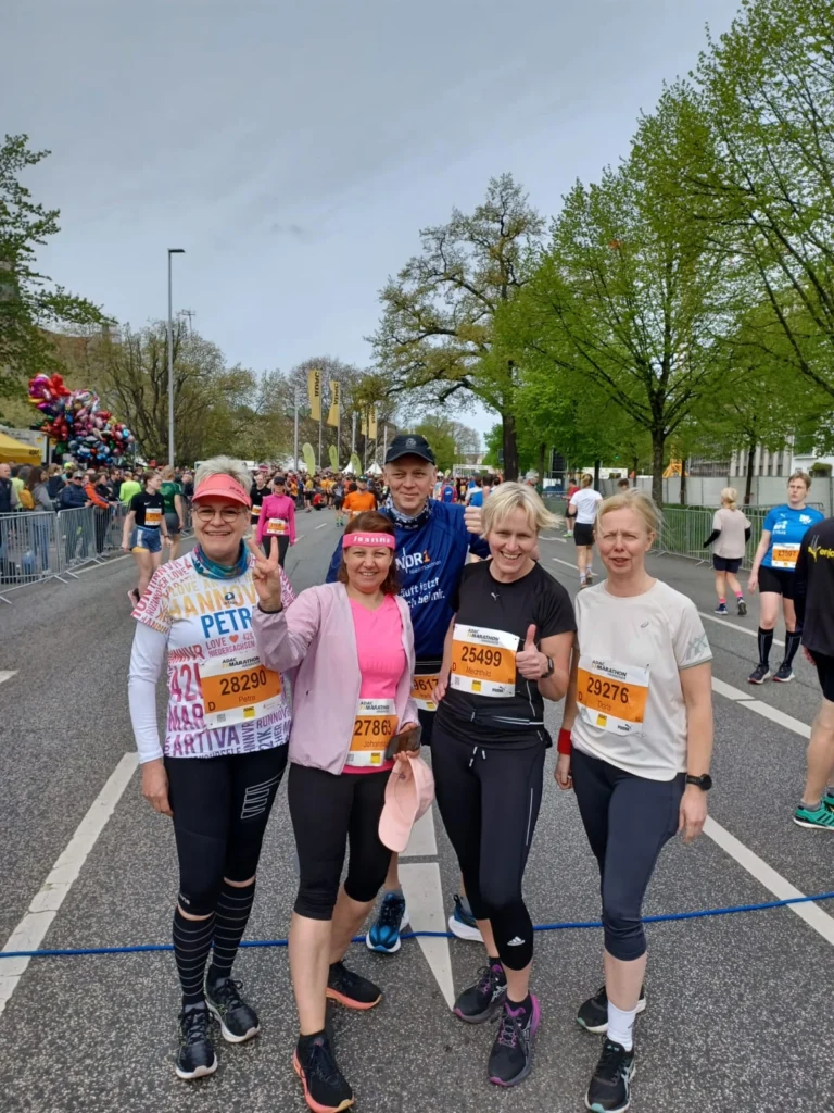 Fünf Teilnehmerinnen und Teilnehmer eines Freundschaftslaufs stehen lächelnd auf der Strecke, umgeben von anderen Läufern und prunkvollen Luftballons. Die Veranstaltung findet an einem sonnigen Tag in einer städtischen Umgebung mit Bäumen im Hintergrund statt. Alle tragen sportliche Outfits und Startnummern.