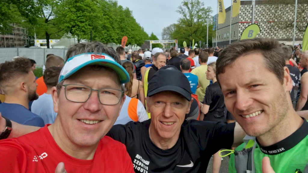 Drei Männer posieren für ein Selfie vor dem Start eines Lauf-Events, umgeben von einer großen Menschenmenge in sportlicher Kleidung, mit Bäumen im Hintergrund und Wettkampf-Banner sichtbar.