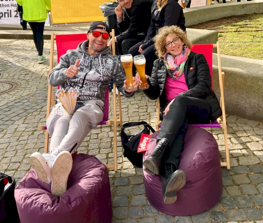 Zwei Personen genießen ein entspanntes Ambiente auf Liegestühlen, halten Biergläser in der Hand und posieren lächelnd. Im Hintergrund sind andere Besucher eines Events zu sehen, was eine gesellige Atmosphäre vermittelt.