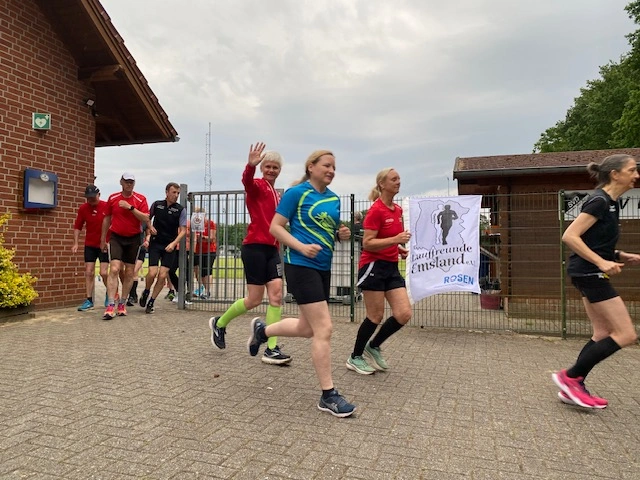 Läufergruppe beim Training vor einem Vereinsgebäude in Deutschland, mit mehreren Sportlern in Laufkleidung, die fröhlich in Richtung Kamera laufen. Im Hintergrund ist ein Banner der Laufgruppe zu sehen.