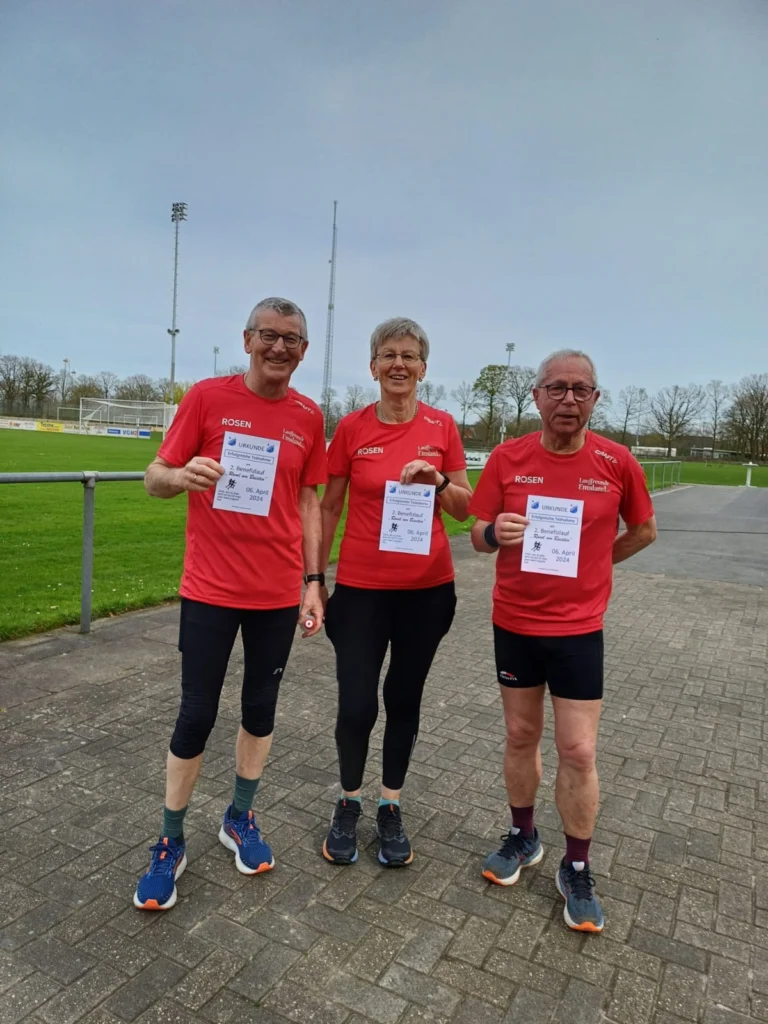 Drei Senioren in roten Laufshirts halten Urkunden für ihre Teilnahme an einem Laufwettbewerb. Die Veranstaltung fand am 6. April 2024 statt, und die Teilnehmer stehen auf einem Sportplatz im Freien.