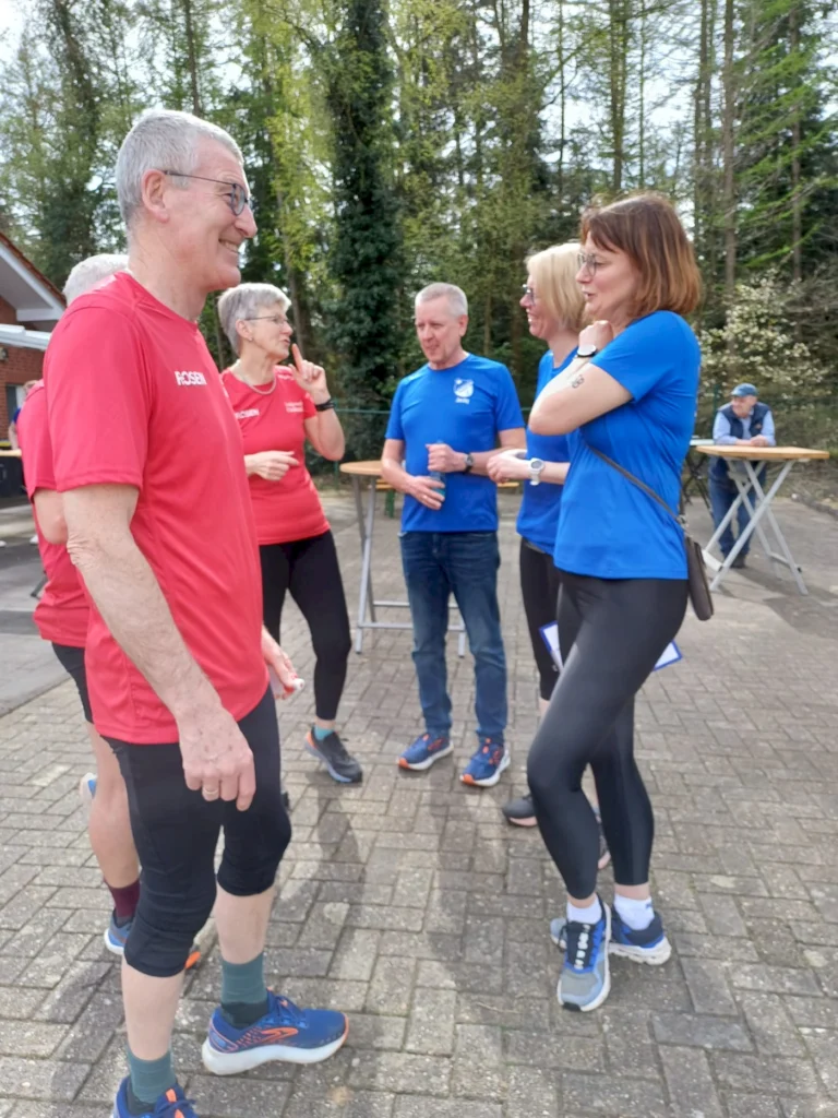 Gruppe von fünf Personen in Sportkleidung, die fröhlich im Freien miteinander sprechen. Zwei Männer tragen rote T-Shirts, während die Frauen in blauen T-Shirts gekleidet sind. Die Szenerie findet in einem parkähnlichen Bereich mit Bäumen im Hintergrund statt.