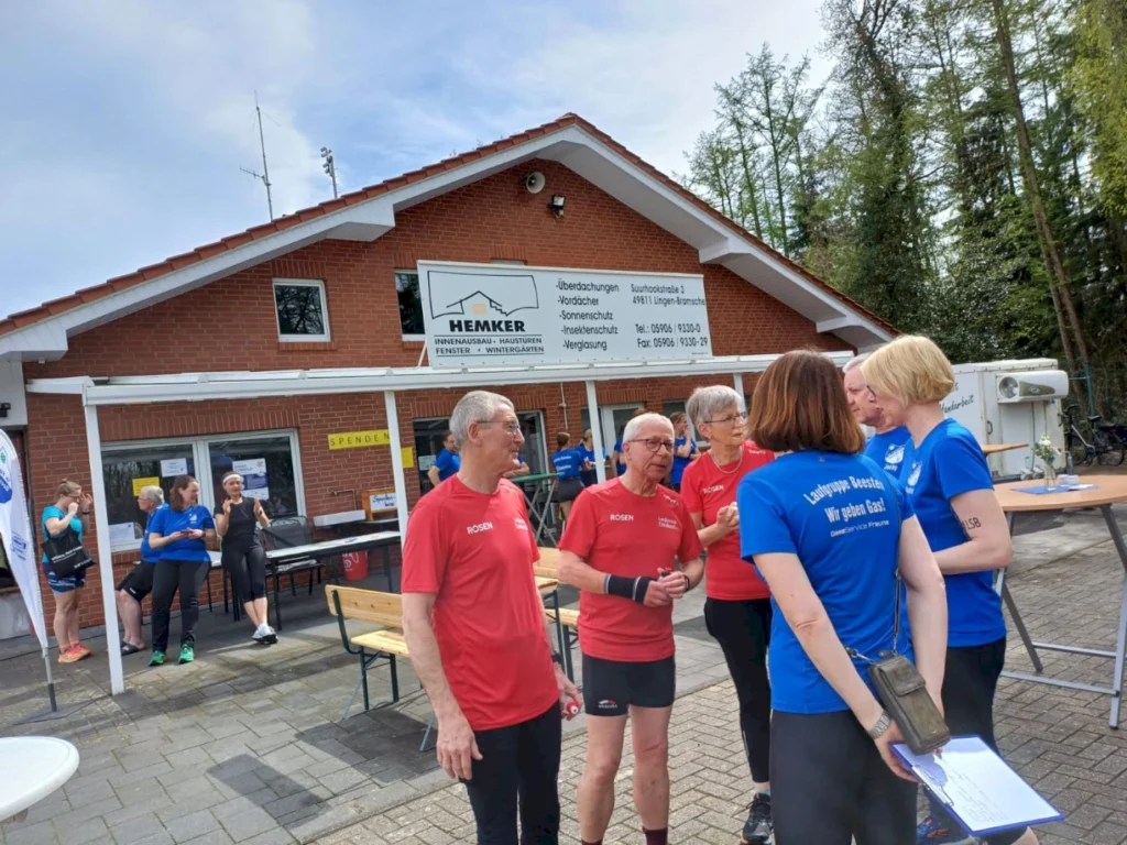 Gruppengespräch von Teilnehmern eines Laufereignisses vor dem Hemker-Gebäude in Lingens-Brammingen, mit mehreren Personen in roten und blauen T-Shirts, die sich austauschen und auf die Veranstaltung vorbereiten.