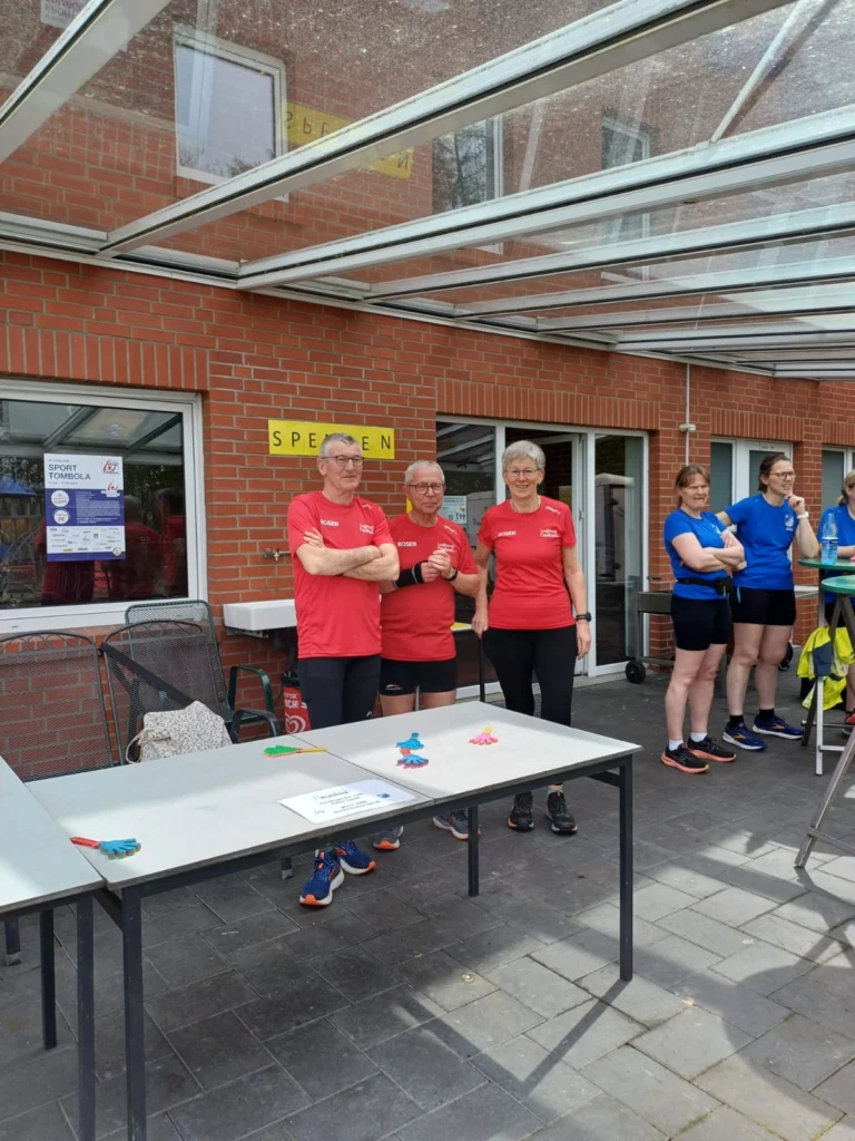 Drei Personen in roten T-Shirts stehen vor einem Tisch im Freien, während sie an einer Sportveranstaltung teilnehmen. Auf dem Tisch liegen bunte Spielzeuge und ein Schild mit Informationen zur Veranstaltung. Im Hintergrund ist ein Gebäude mit Fenstern und einer Überdachung zu sehen.