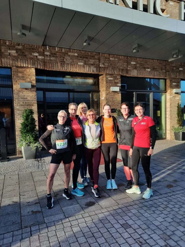 Gruppe von sieben Läuferinnen in Sportkleidung, die vor einem modernen Gebäude stehen, bereit für einen Wettkampf. Die Teilnehmer tragen Startnummern und zeigen eine motivierte Stimmung.