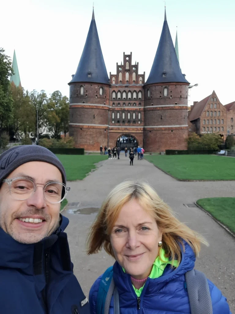 Paar posiert vor dem historischen Holstentor in Lübeck, Deutschland, umgeben von einer grünen Parklandschaft und anderen Besuchern.