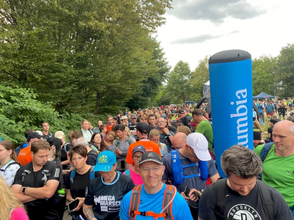 Große Menschenmenge bei einem Outdoor-Event mit Teilnehmern in Sportkleidung, umgeben von Bäumen und einem blauen Werbebanner von Columbia.