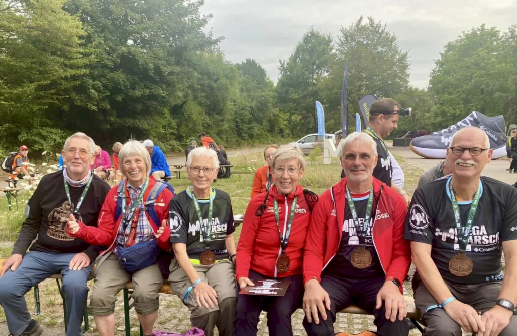 Gruppe von sechs Personen in Sportbekleidung mit Medaillen, die nach einem Wettkampf im Freien auf Bänken sitzen. Im Hintergrund sind weitere Teilnehmer und Banner sichtbar.