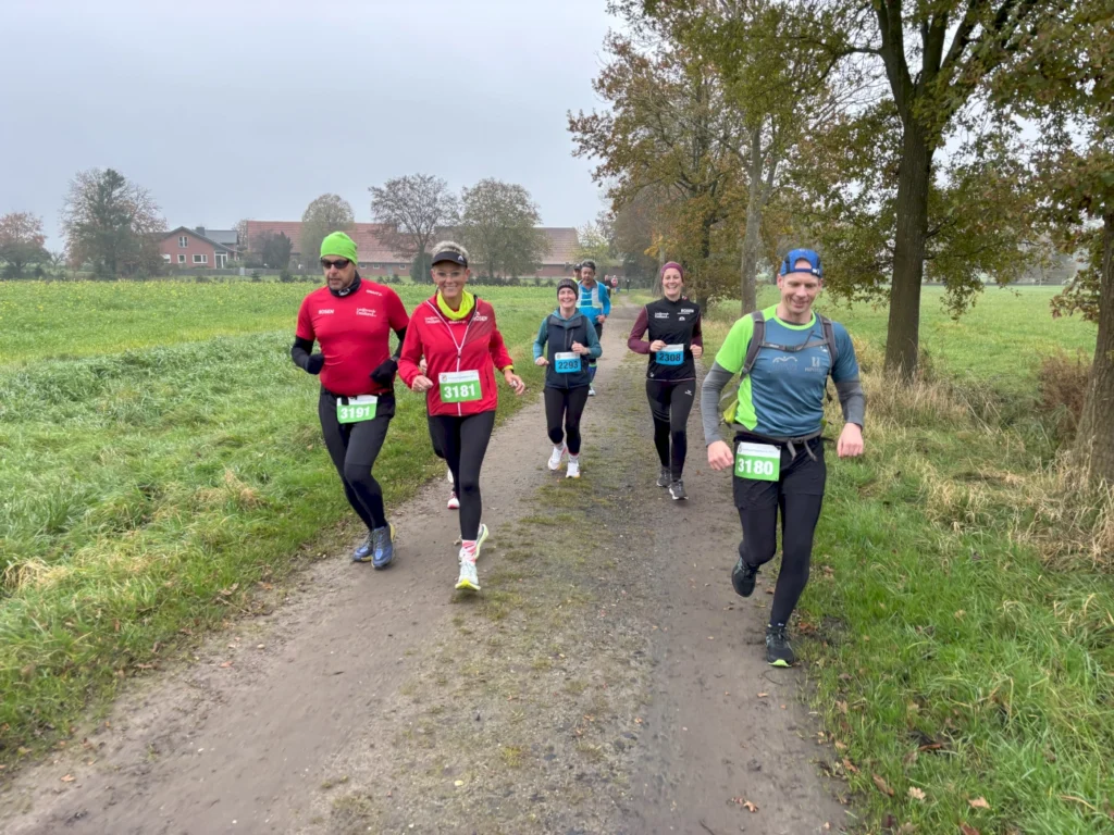 Gruppe von Läufern bei einem Volkslauf auf einem Feldweg, umgeben von grünen Wiesen und Bäumen, mit unterschiedlichen Laufkleidungen und Startnummern.