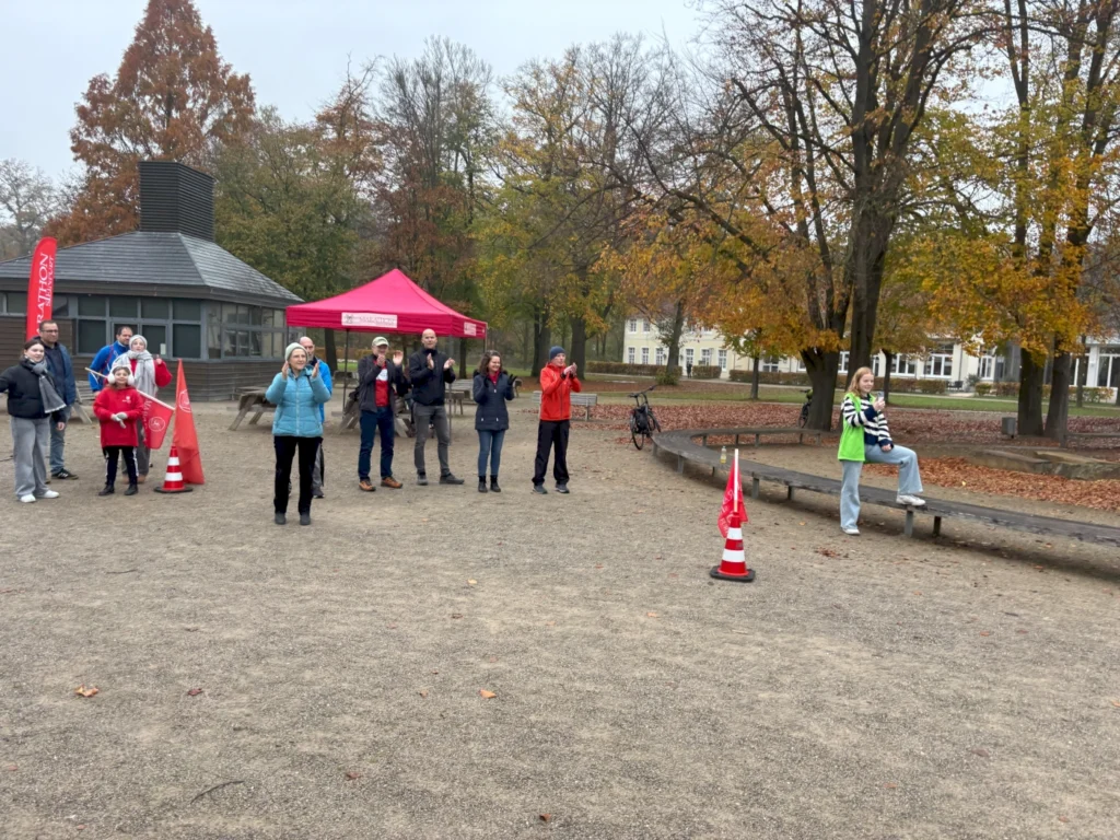 Versammlung von Menschen im Park bei trübem Wetter, einige tragen Winterkleidung, während sie applaudieren und Zuschauer sind. Im Hintergrund steht ein rotes Zelt und herbstliche Bäume sind zu sehen.