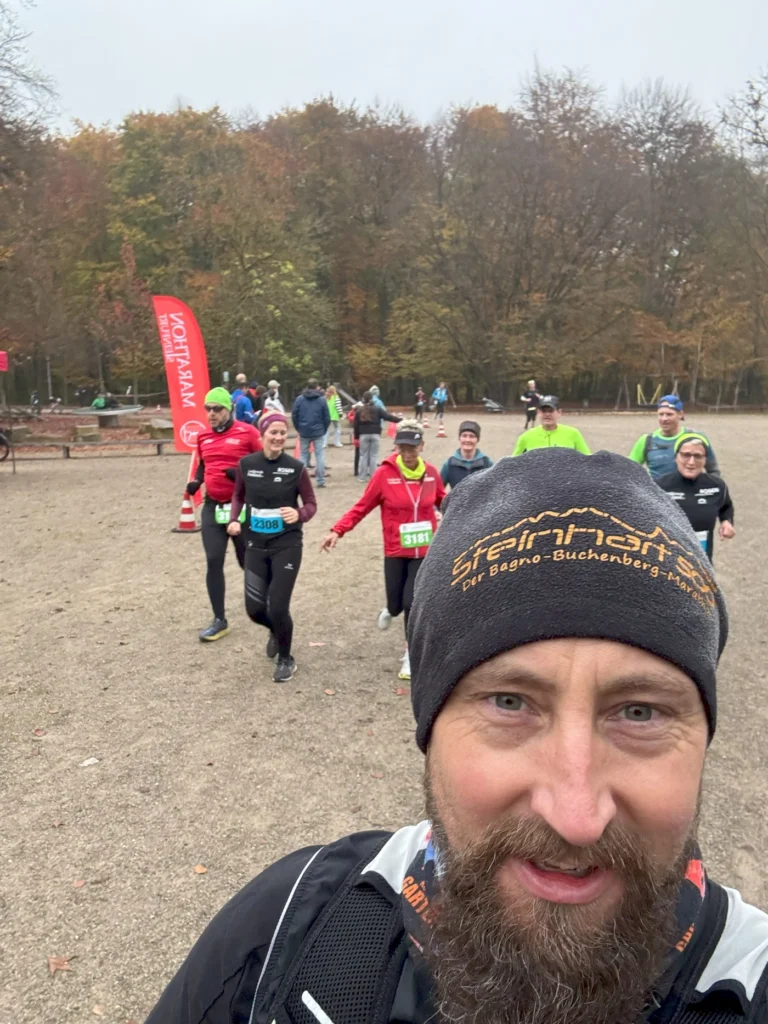 Läufer beim Wettkampf im Wald mit herbstlichen Bäumen im Hintergrund. Der Fokus liegt auf einem Mann mit einem Bart und einer Mütze, während andere Teilnehmer im Hintergrund vorbeilaufen. Ideal für Beiträge über Laufveranstaltungen und Outdoor-Sportarten.
