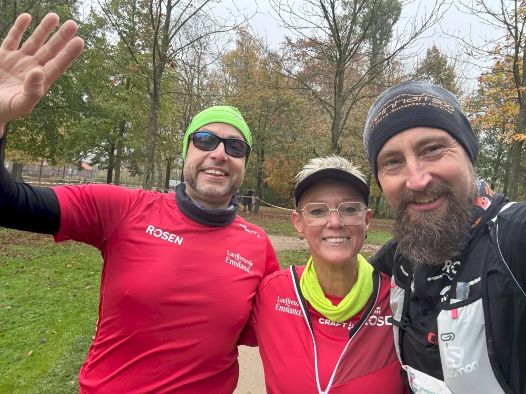 Drei sportliche Läufer posieren lächelnd in einem Park während eines Lauf-Events. Sie tragen rote T-Shirts mit dem Logo "RÖSEN" und andere Laufbekleidung, umgeben von herbstlichen Bäumen.