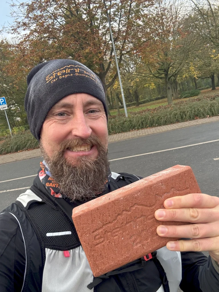 Läufer mit Bart hält einen Ziegelstein in der Hand, trägt eine Mütze mit dem Logo eines Outdoor-Unternehmens und steht in einem Park mit herbstlichen Bäumen im Hintergrund.