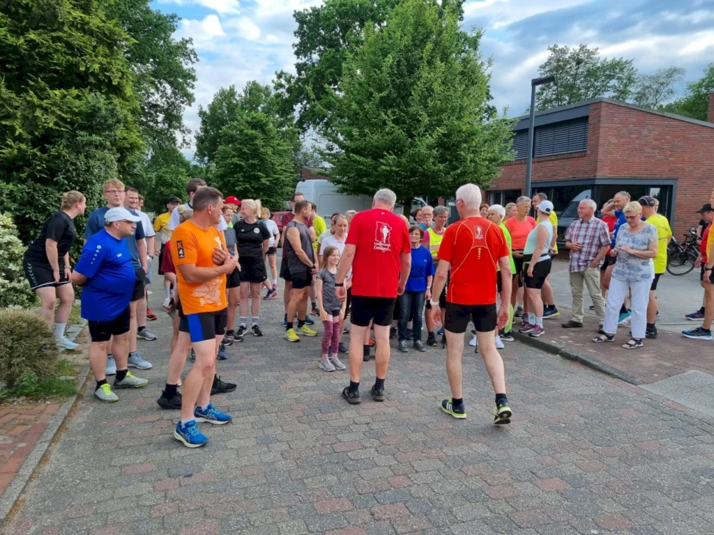Gruppierung von Läufern und Zuschauern bei einem Laufereignis im Freien, umgeben von Bäumen und Gebäuden. Die Teilnehmer tragen sportliche Kleidung und bereiten sich auf den Start vor.