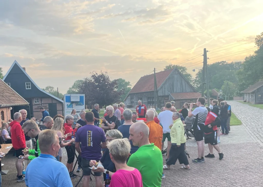 Menschenansammlung bei einem Outdoor-Event in ländlicher Umgebung, mit Teilnehmern, die Snacks genießen und sich unterhalten, während die Sonne aufgeht. Historische Gebäude im Hintergrund, die eine gemütliche Atmosphäre schaffen.