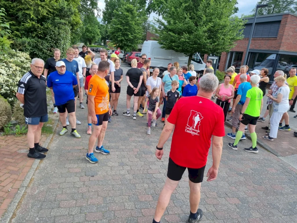 Gruppe von Läufern in verschiedenen Sportoutfits versammelt sich an einem sonnigen Tag auf einem gepflasterten Weg, um an einem Laufereignis in Deutschland teilzunehmen. Im Hintergrund sind Bäume und Fahrzeuge zu sehen.