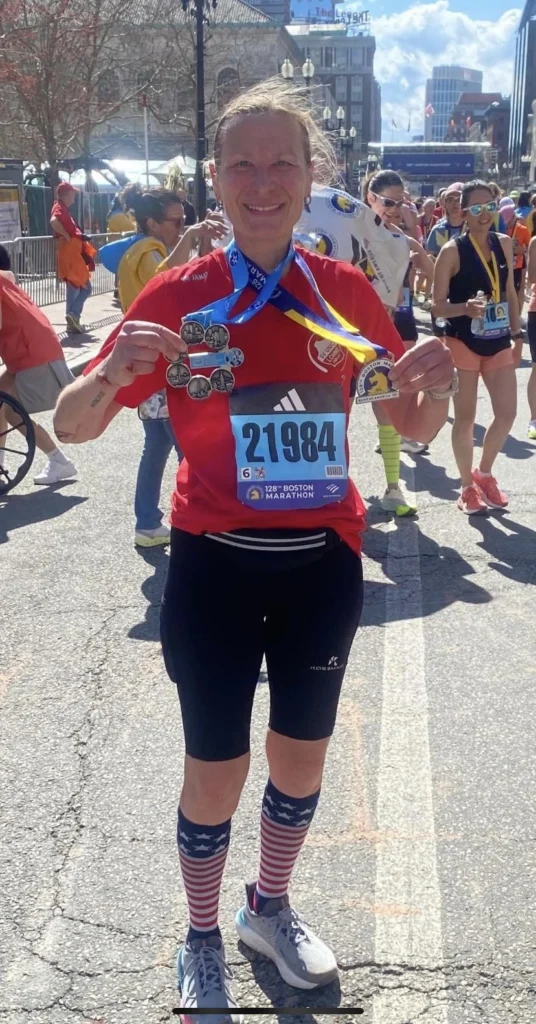 Läuferin beim Boston-Marathon 2024, trägt Medaillen und ein rotes Shirt mit der Startnummer 21984, im Hintergrund andere Teilnehmer und eine sonnige Atmosphäre.