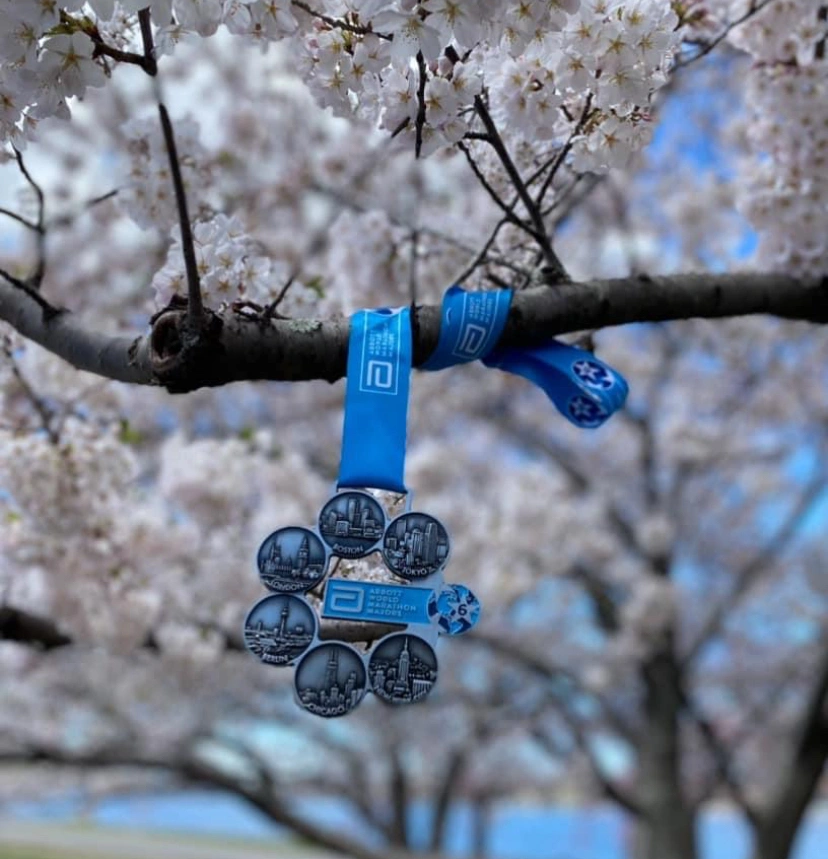 Medaille mit mehreren Medaillenringen, die an einem Baumzweig mit blühenden Kirschblüten hängen, im Hintergrund blauer Himmel. Die Medaille repräsentiert verschiedene Städte, darunter Boston und Tokyo.