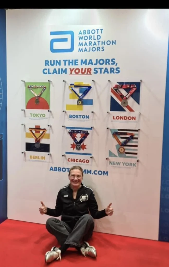 Person sitzt lächelnd auf dem Boden vor dem Banner der Abbott World Marathon Majors, das Medaillen von sechs großen Marathons zeigt: Tokyo, Boston, London, Berlin, Chicago und New York.