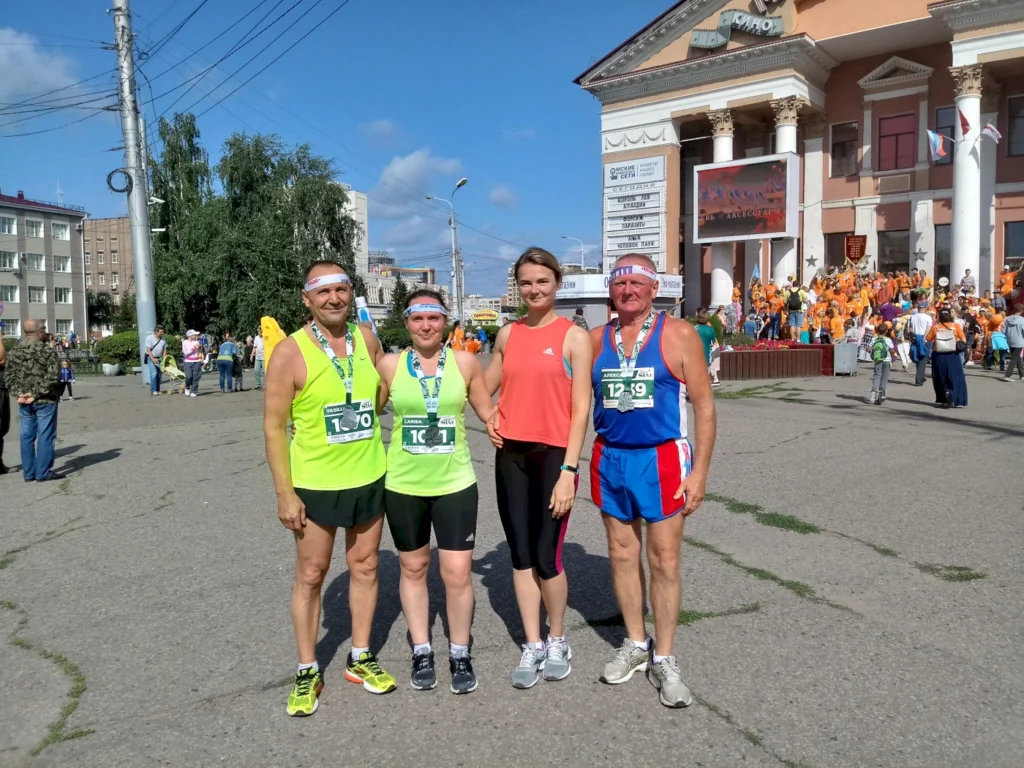 Vier Läufer nach einem Wettkampf, alle mit Medaillen um den Hals, stehen vor einem Gebäude mit festlicher Atmosphäre. Sie tragen Sportbekleidung in verschiedenen Farben und posieren lächelnd für das Foto, während im Hintergrund eine Menschenmenge feiert.