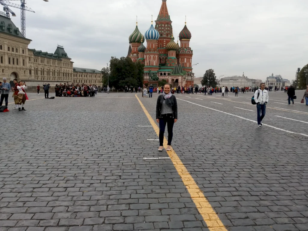 Frau steht auf dem Roten Platz in Moskau, mit der Basilius-Kathedrale im Hintergrund. Die Umgebung ist belebt, mit Touristen und Einheimischen, die die Sehenswürdigkeiten erkunden. Der Himmel ist bewölkt, und die Pflastersteine des Platzes sind deutlich sichtbar.