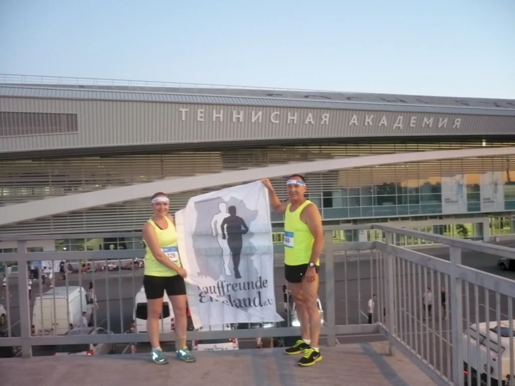 Zwei Läufer in sportlicher Kleidung halten ein Banner mit der Aufschrift "Lauffreunde Deutschland" vor einem modernen Gebäude der Tennisakademie. Die Szene spielt sich während einer Laufveranstaltung ab, die in den frühen Morgenstunden stattfindet.