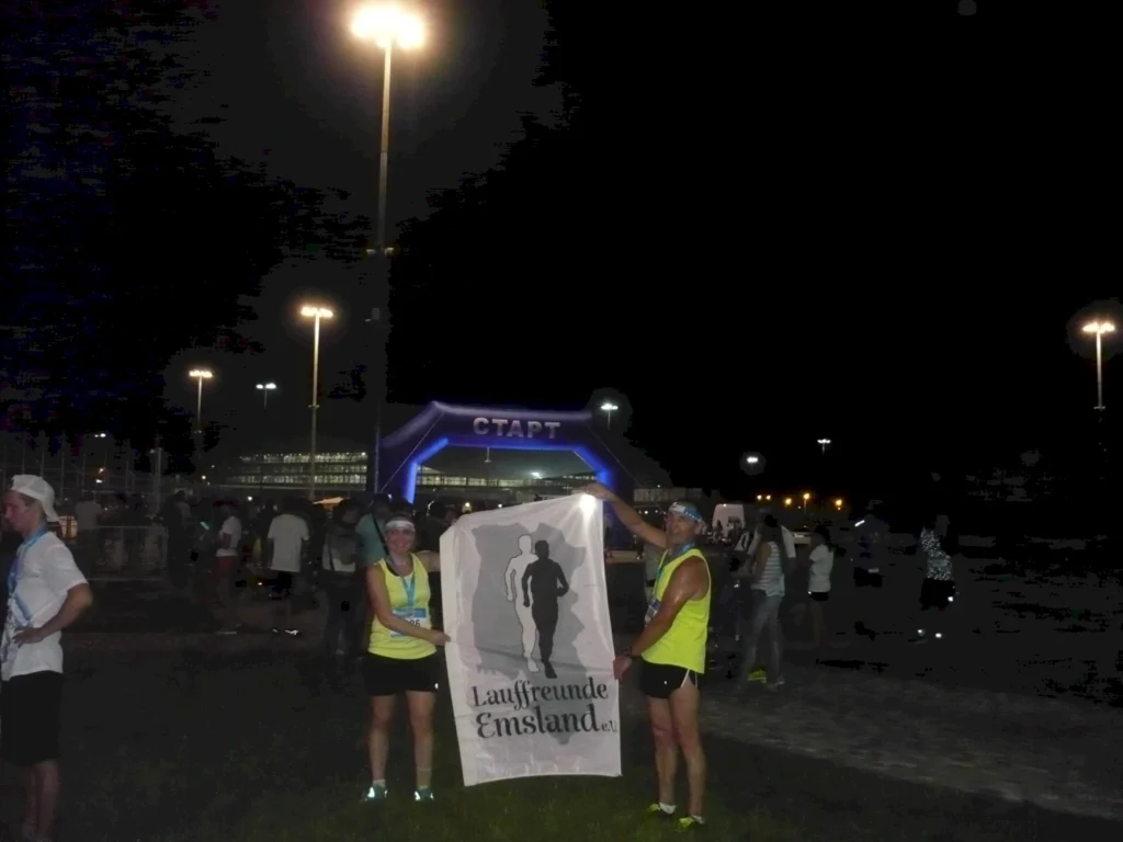 Zwei Läufer der Laufgruppe Emsland halten ein Banner bei Nacht vor dem Startbereich eines Laufevents. Beleuchtung und Teilnehmer im Hintergrund.