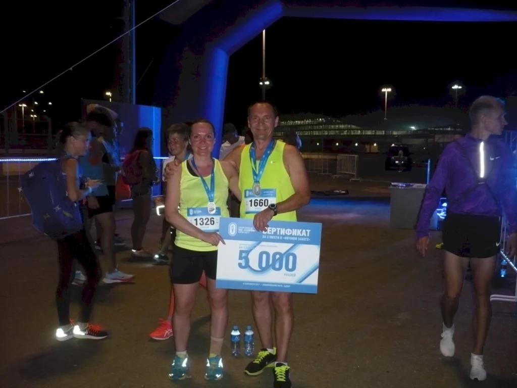 Läufer stehen bei Nacht nach einem Wettkampf zusammen, halten Medaillen und ein Zertifikat über 3.000 Rubel. Sie tragen sportliche Kleidung und lächeln in die Kamera, während im Hintergrund weitere Teilnehmer zu sehen sind.