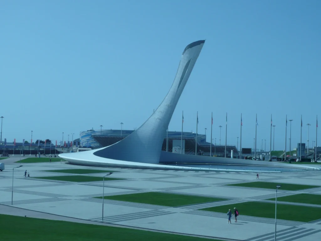 Architektonisches Wahrzeichen in Sotschi mit futuristischem Design, umgeben von einer weitläufigen Platzlandschaft und Flaggen. Im Hintergrund ist das Stadion zu sehen.