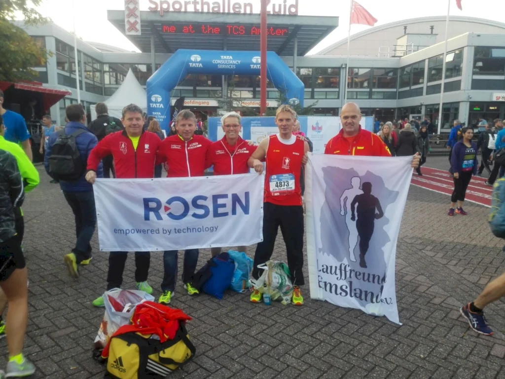 Teilnehmer des TCS Marathons stehen vor dem Sporthallen Süd, halten Banner von ROSEN und dem Verein Lauffreund Emsland e.V. in der Hand. Die Veranstaltung zieht zahlreiche Läufer und Zuschauer an, während die Atmosphäre von Vorfreude geprägt ist.