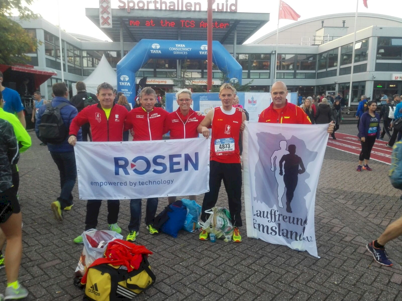 Teilnehmer des TCS Marathons stehen vor dem Sporthallen Süd, halten Banner von ROSEN und dem Verein Lauffreund Emsland e.V. in der Hand. Die Veranstaltung zieht zahlreiche Läufer und Zuschauer an, während die Atmosphäre von Vorfreude geprägt ist.