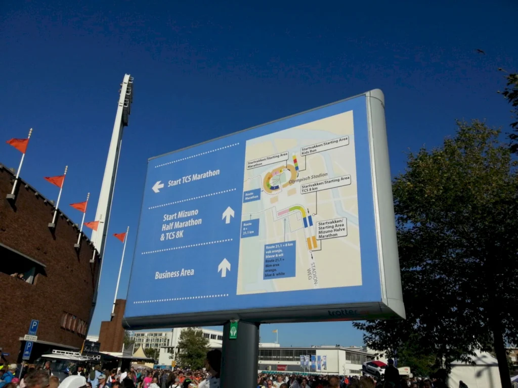 Informationsschild zum TCS Marathon und Mizuno Halbmarathon mit Startpunkten und Streckenplan, Olympiastadion im Hintergrund, blauer Himmel und Menschenmenge.