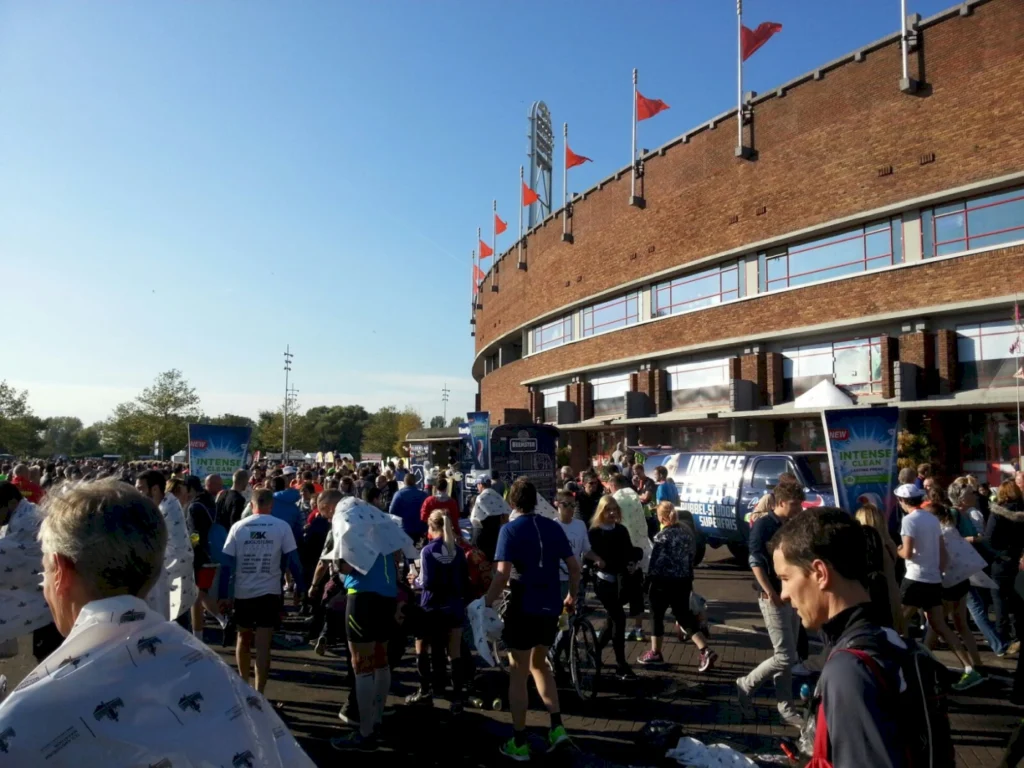Menschenmenge vor einem Stadion während eines Lauf-Events, mit Teilnehmern in Sportkleidung und Decken, bei klarem Himmel und sonnigem Wetter.