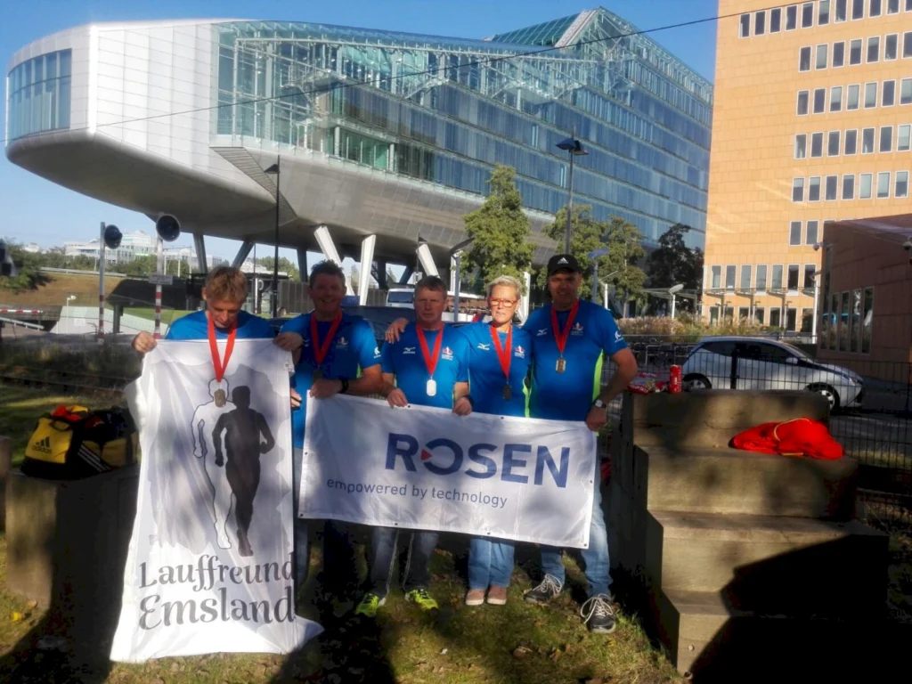 Gruppe von Läufern in blauen T-Shirts mit Medaillen, die ein Banner von "Lauffreund Emsland" und ein Firmenbanner halten, vor einem modernen Gebäude.