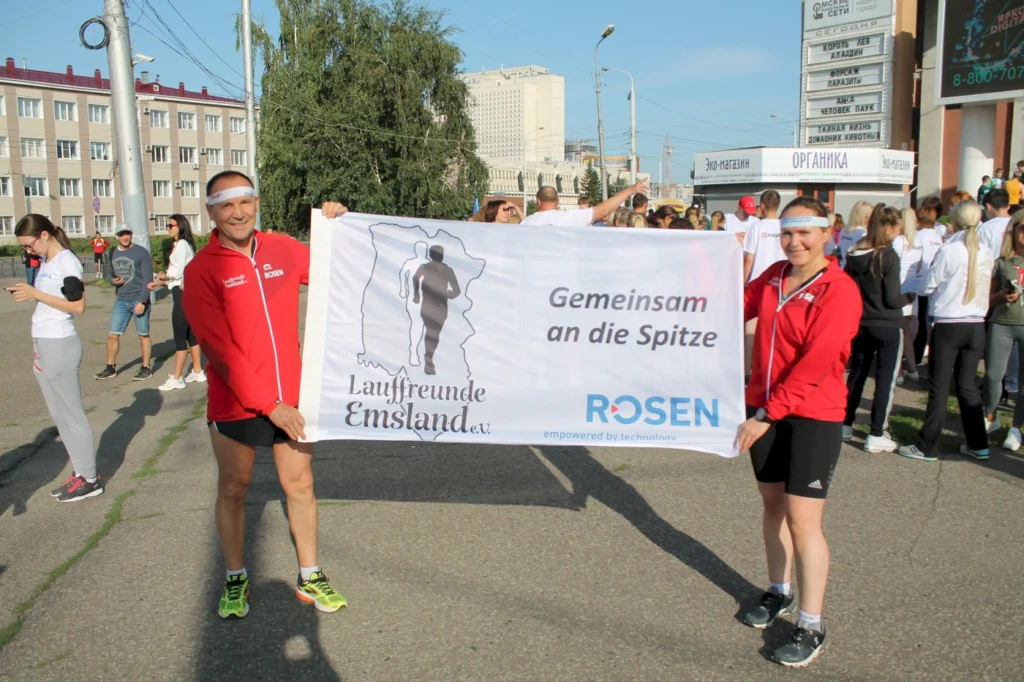 Zwei Läufer der Lauffreunde Emsland halten ein Banner mit der Aufschrift „Gemeinsam an die Spitze“ während einer Laufveranstaltung, umgeben von weiteren Teilnehmern in sportlicher Kleidung.