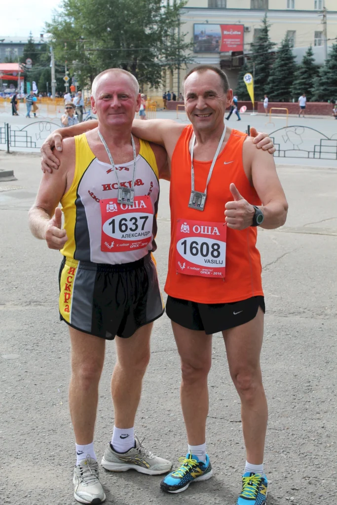 Zwei männliche Läufer nach einem Wettkampf, einer in einem gelben und grauen Trikot, der andere in einem orangefarbenen Trikot, beide mit Medaillen um den Hals und lächelnd mit Daumen hoch. Im Hintergrund sind Zuschauer und eine Veranstaltungskulisse zu sehen.