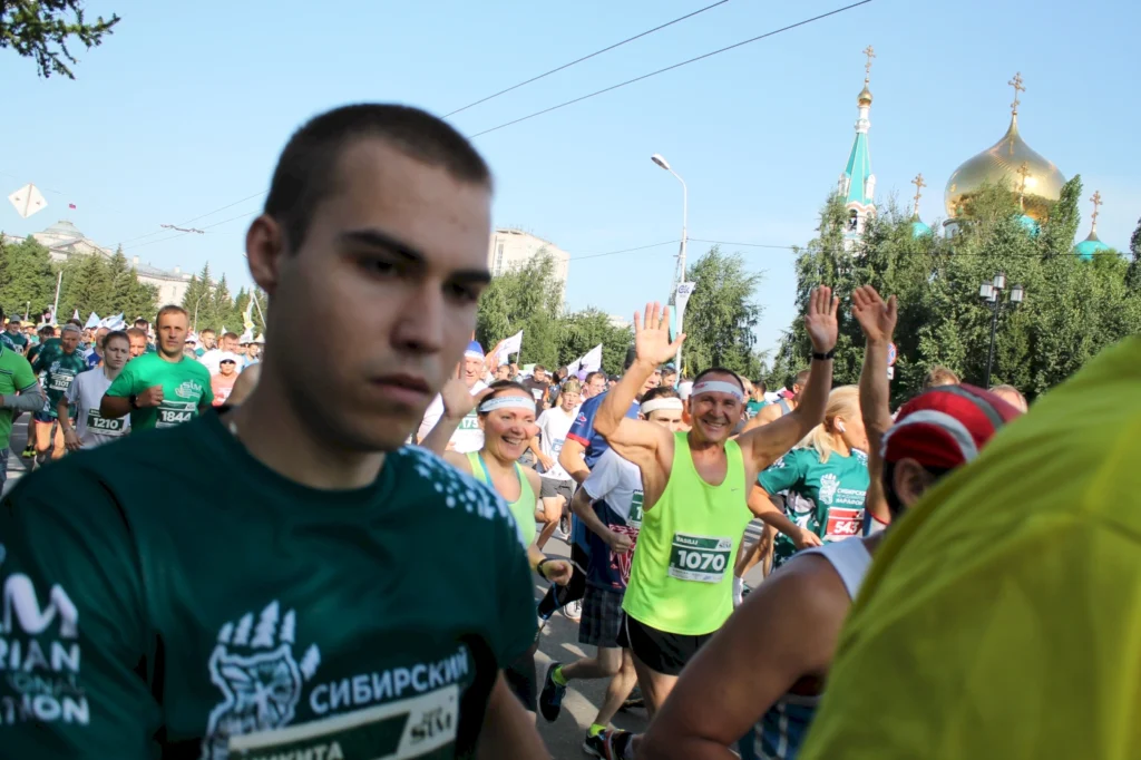 Läufer bei einem Wettlauf in einer Stadt mit grünen Bäumen und goldenen Kuppeln im Hintergrund. Der Fokus liegt auf einem jungen Mann im Vordergrund und fröhlichen Teilnehmern, die sich an der Veranstaltung beteiligen.