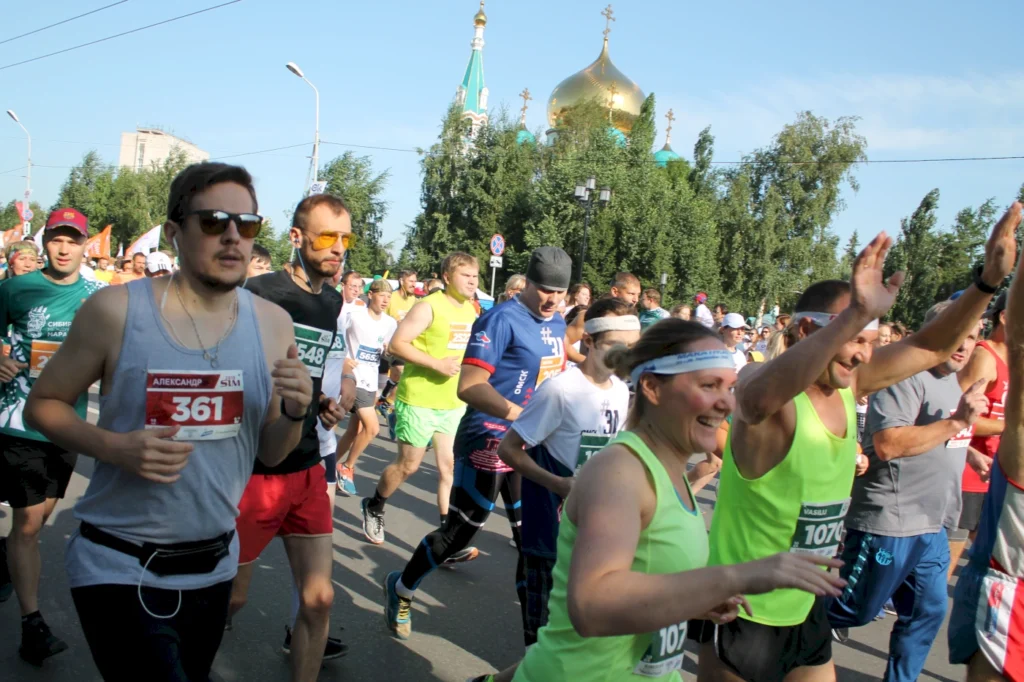 Teilnehmer eines Straßenlaufs mit verschiedenen Sportoutfits, darunter einige mit Startnummern, laufen unter einem klaren Himmel in einer Stadt, im Hintergrund sind das grüne Kupolengebäude und Bäume sichtbar.